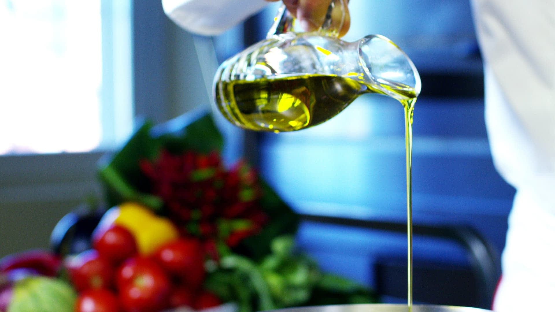 A person pouring olive oil from a glass bottle into a metal bowl in a kitchen setting. - Olive Oil Times