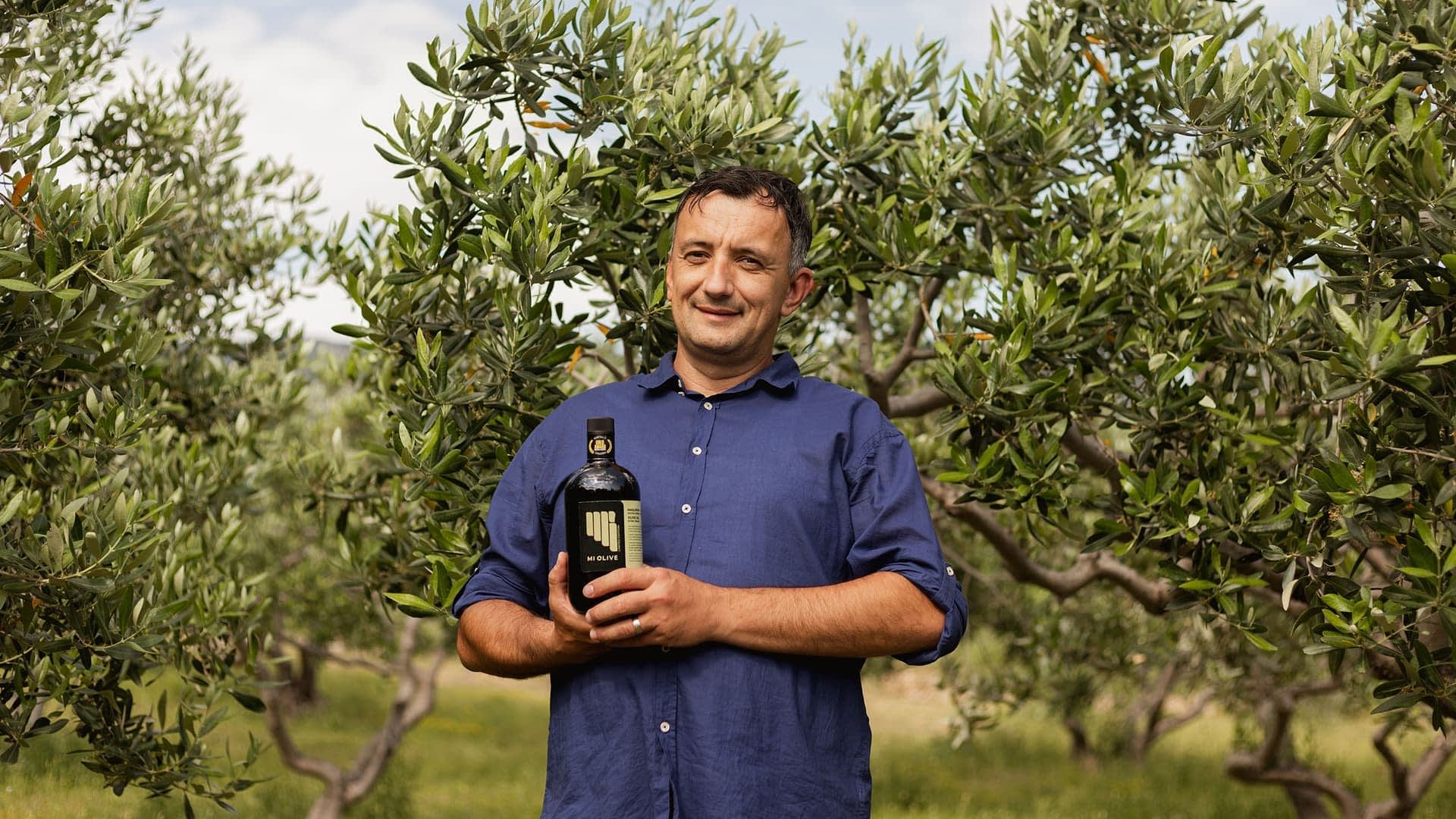 Man standing in an olive grove holding a bottle of olive oil with olive trees in the background. - Olive Oil Times