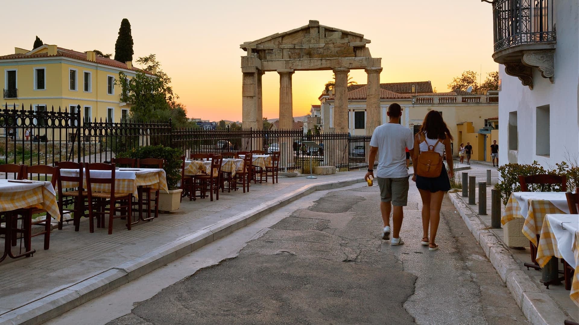 A couple walking along a street near ancient ruins at sunset, with outdoor dining tables visible. - Olive Oil Times