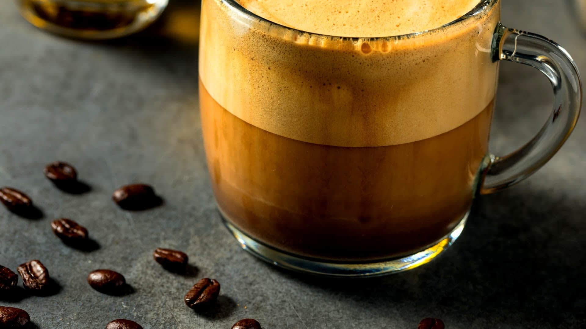 A glass mug filled with a layered coffee beverage, featuring a frothy top and dark liquid below. - Olive Oil Times