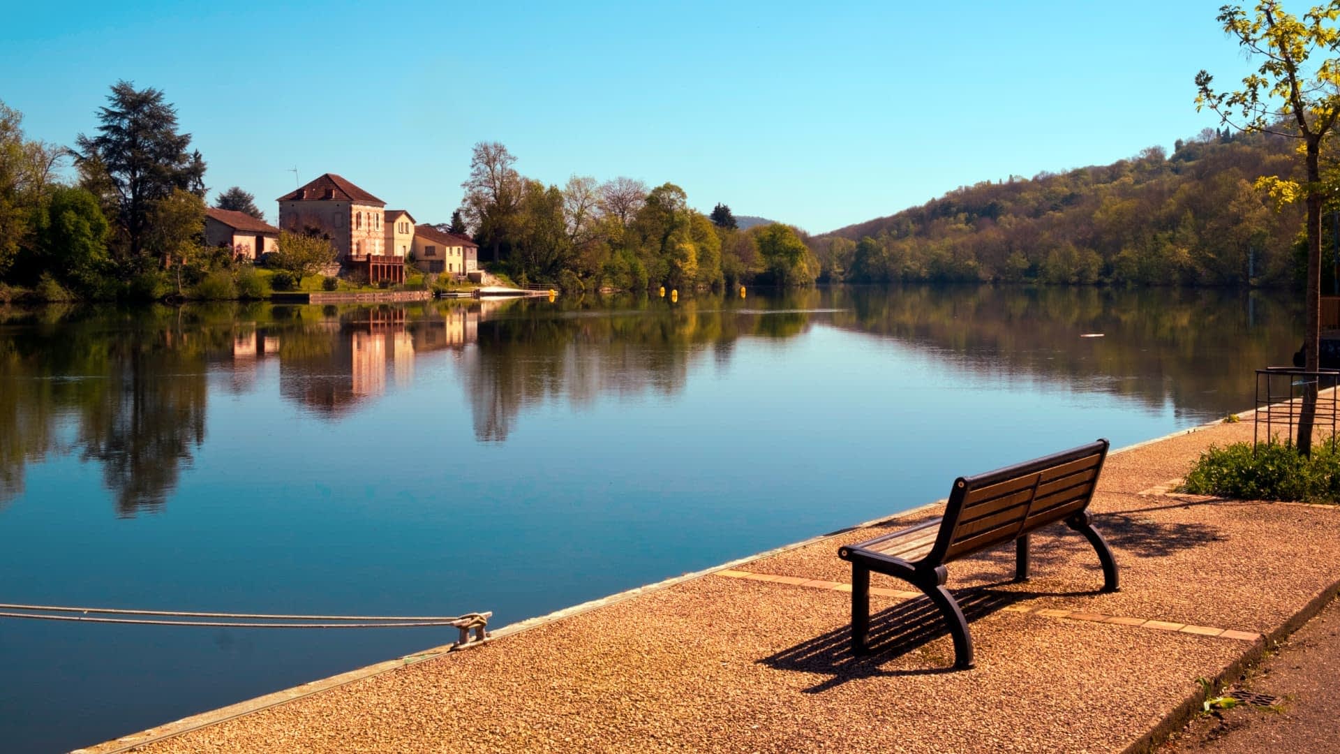 A wooden bench positioned along the edge of a calm river reflecting trees and buildings in the water. - Olive Oil Times