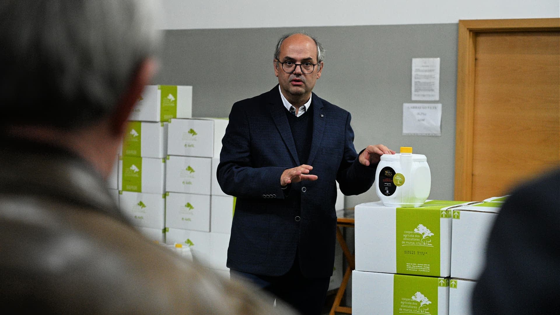 A man in a suit presenting a container of olive oil in a warehouse setting. - Olive Oil Times