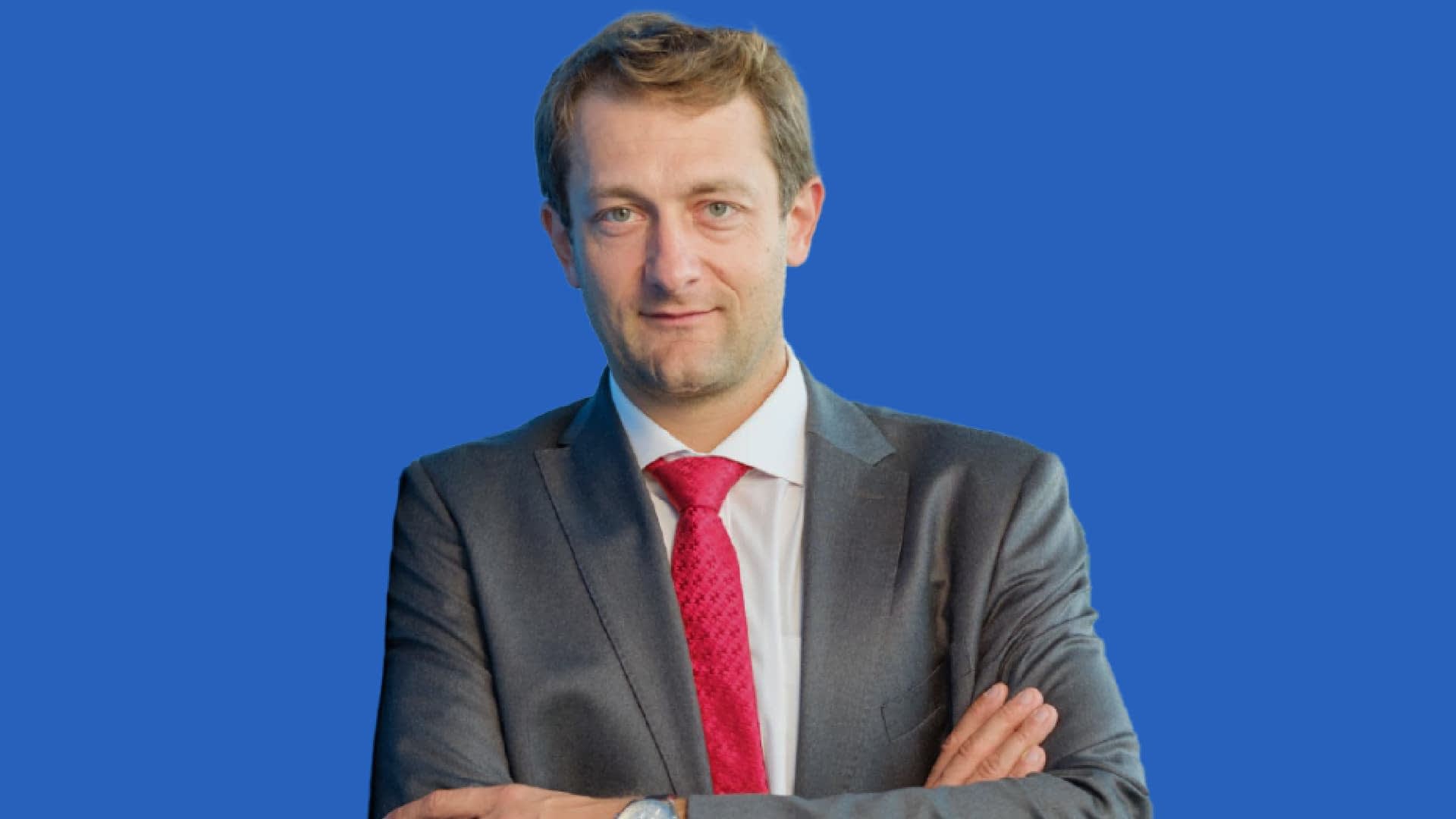 Man wearing a gray suit and a red tie, standing with arms crossed against a blue background. - Olive Oil Times