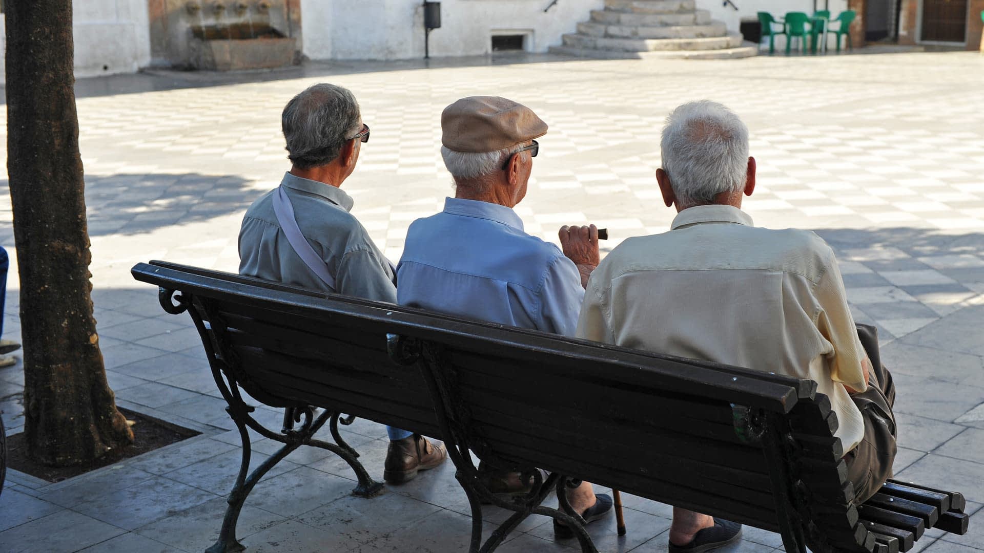 Three elderly men sitting on a bench, viewed from behind, in a public square. - Olive Oil Times