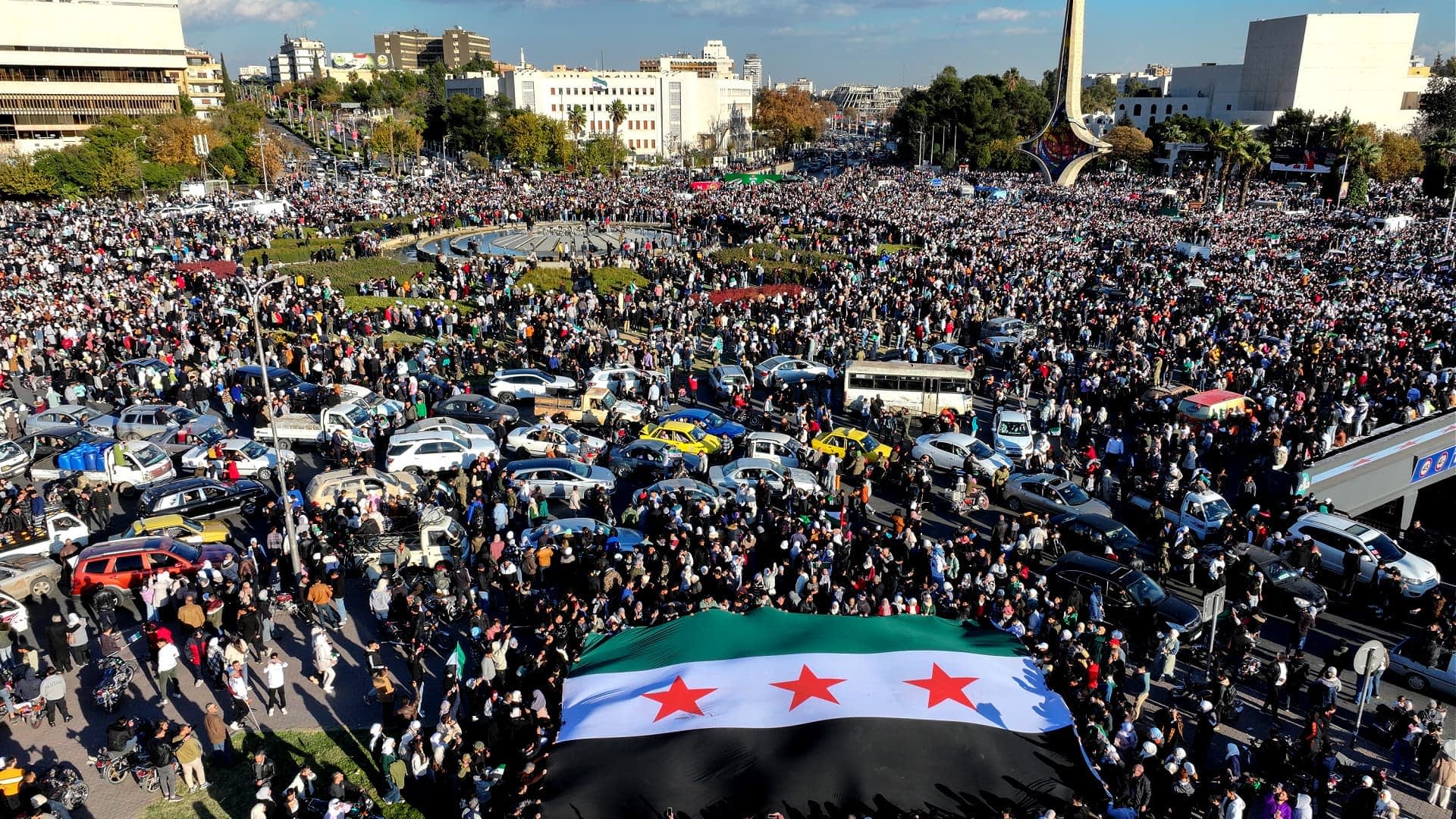 A large crowd gathered in a public square holding a Syrian flag with three stars. - Olive Oil Times