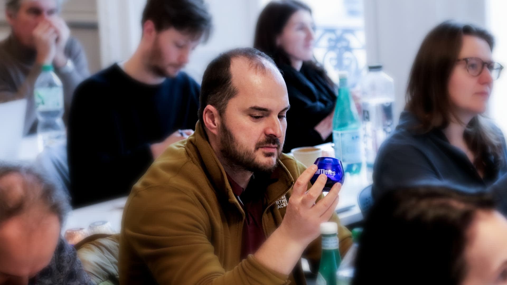 Man examining a blue olive oil bottle while seated in a seminar with other attendees in the background. - Olive Oil Times
