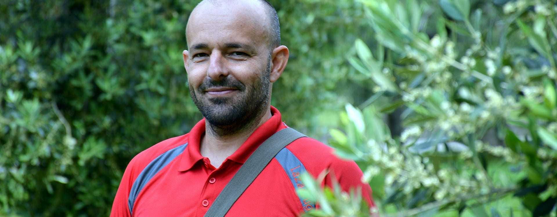 Un homme portant un polo rouge debout parmi les oliviers avec un léger sourire. - Olive Oil Times
