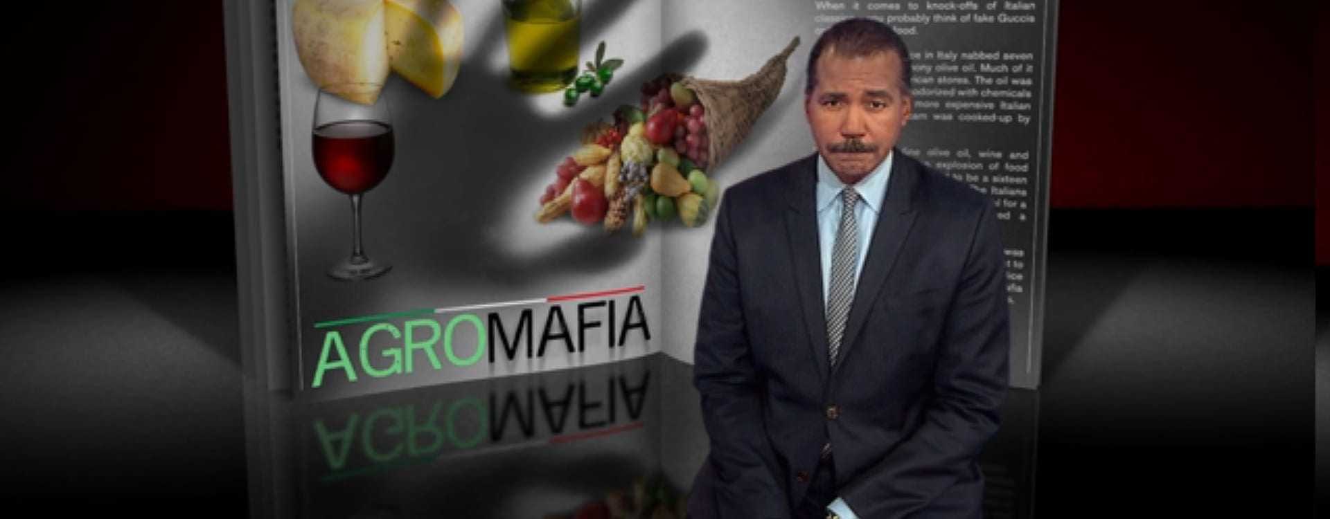 A man in a suit standing next to a graphic titled 'Agro Mafia' with food items and a glass of wine. - Olive Oil Times