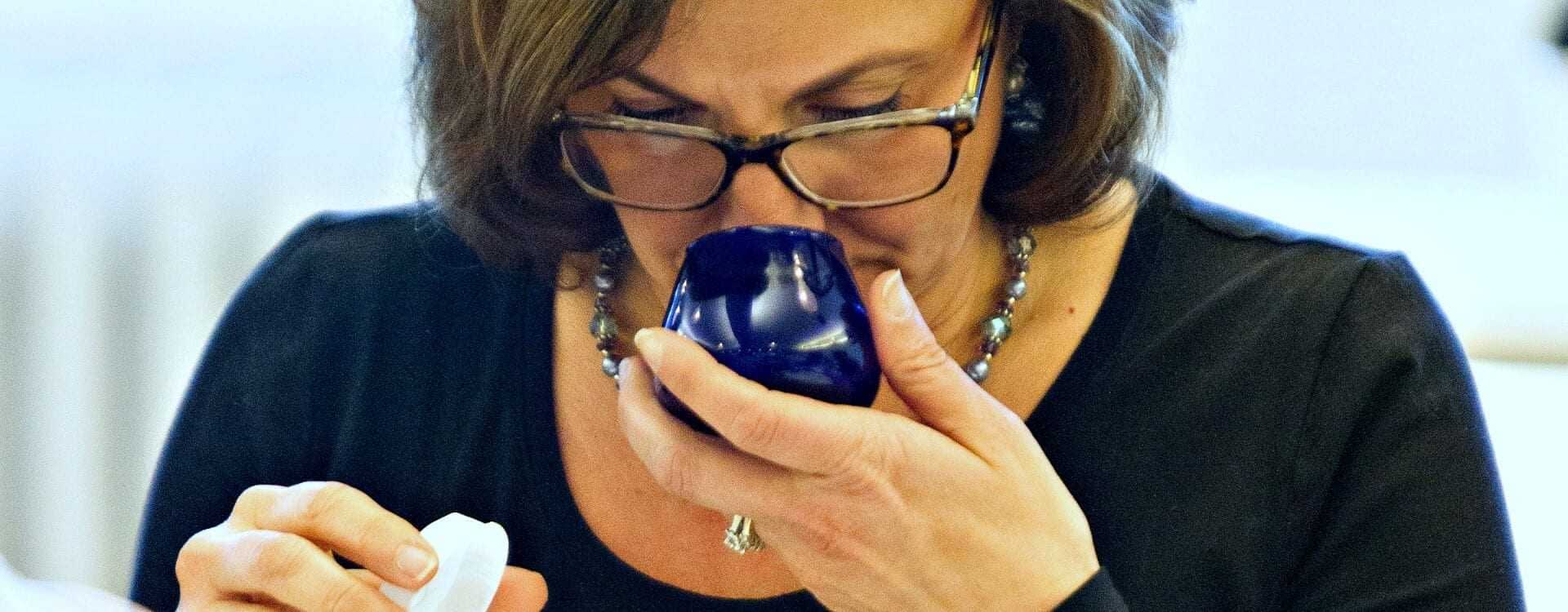 Woman with glasses smelling olive oil from a blue tasting cup while holding a small container. - Olive Oil Times