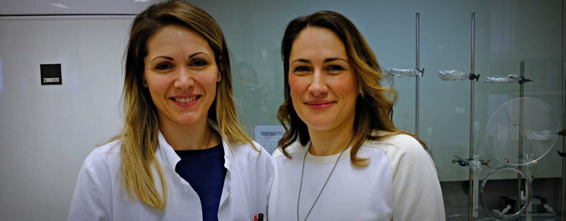 Two women smiling in a laboratory environment, one wearing a lab coat and the other in a white sweater. - Olive Oil Times
