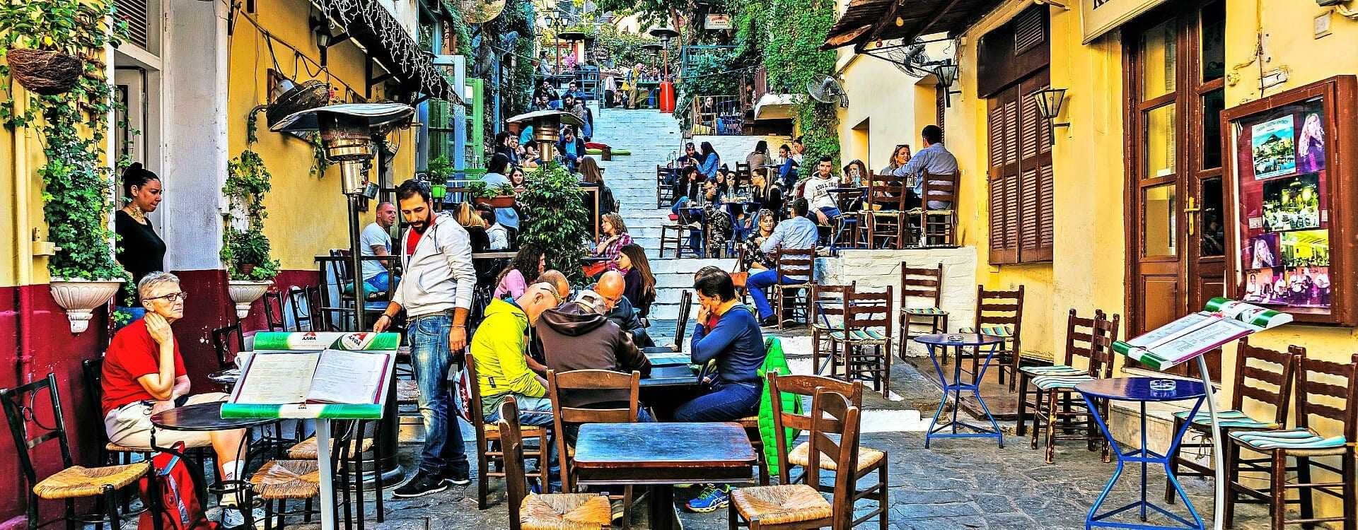 Outdoor dining area featuring wooden tables and chairs in a bustling setting with patrons. - Olive Oil Times