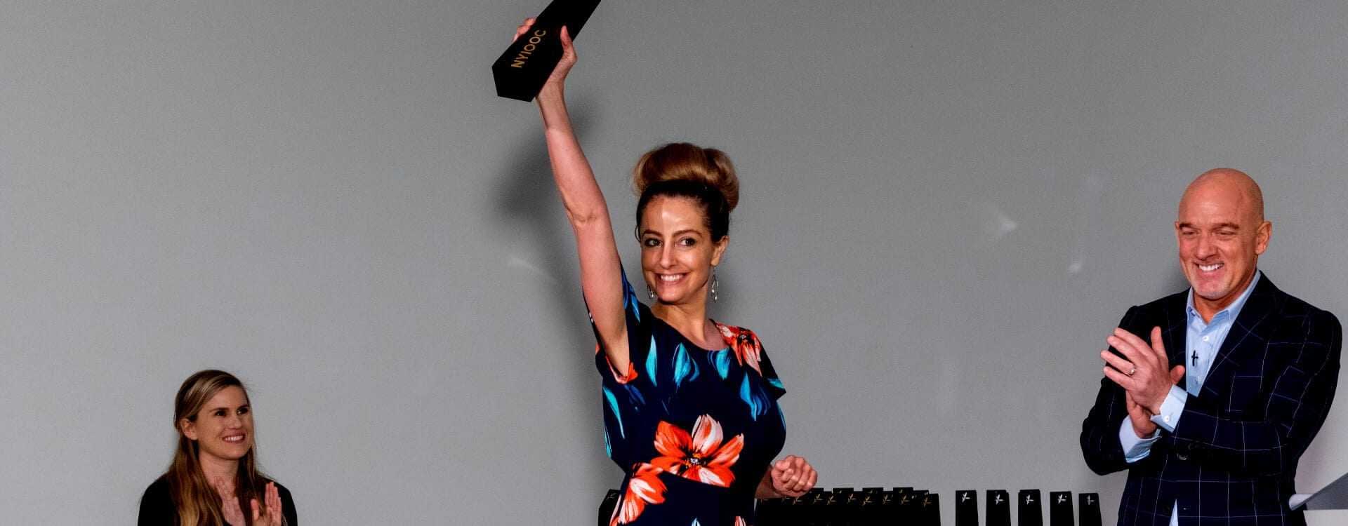 Woman in a floral dress holding an award above her head during an event with two people clapping in the background. - Olive Oil Times