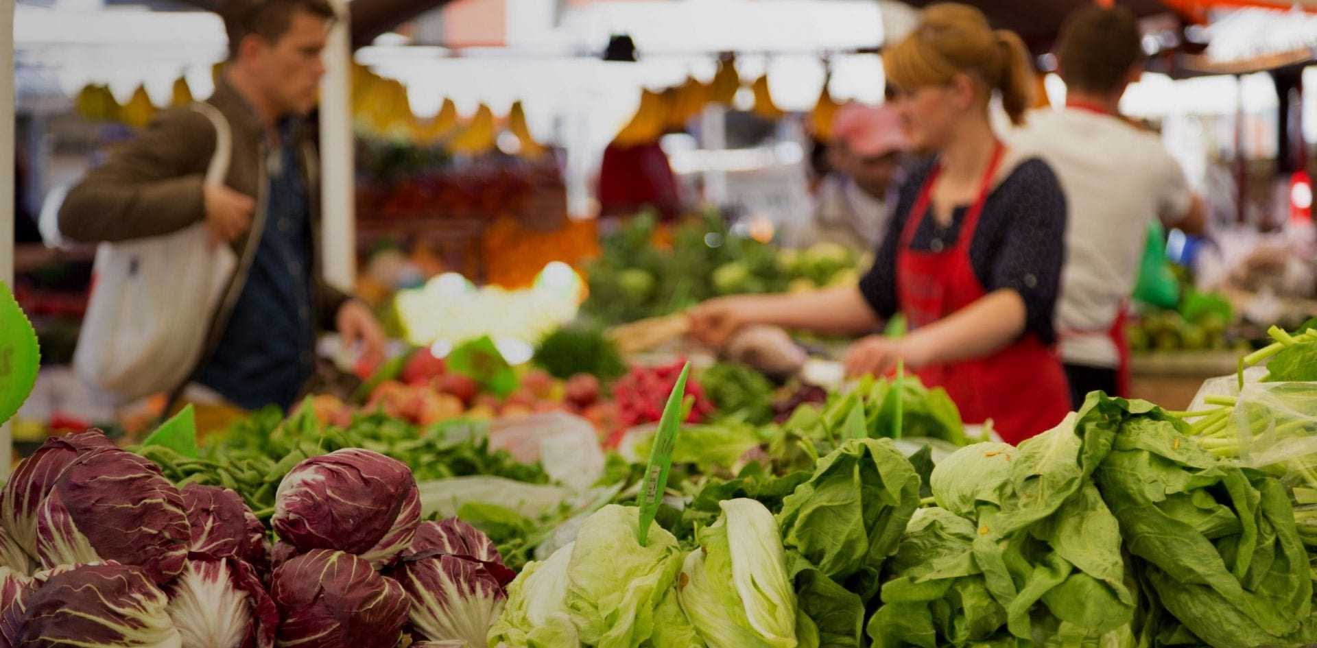 Fresh vegetables including lettuce and radicchio displayed at a market stall with shoppers in the background. - Olive Oil Times
