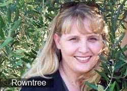 A woman with blonde hair smiling while standing among green leaves and branches. - Olive Oil Times