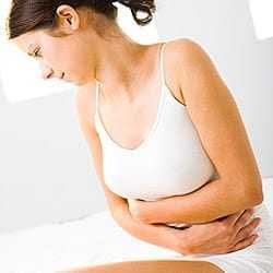 Woman in a white tank top sitting on a bed, holding her stomach with a pained expression. - Olive Oil Times