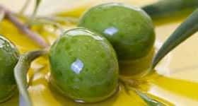 Tri zelene olive počivajo v bazenu oljčnega olja z listi v bližini. - Olive Oil Times