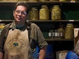 Man wearing an apron standing in an olive shop with jars of olives on shelves behind him. - Olive Oil Times