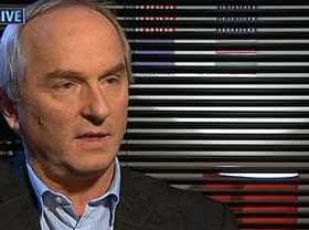 Man with gray hair wearing a dark suit and light shirt during an interview against a striped backdrop. - Olive Oil Times