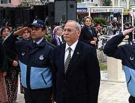 Two police officers saluting alongside a man in a suit at a public event with a crowd in the background. - Olive Oil Times