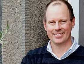 Man with short hair wearing a navy blue sweater, smiling in front of a textured wall. - Olive Oil Times