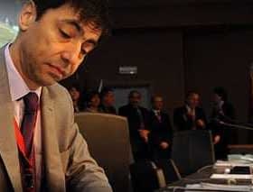 A man in a suit signing documents at a conference table with a serious expression. - Olive Oil Times