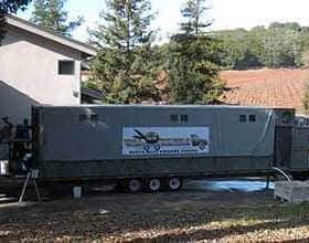 Mobile olive oil processing unit parked next to a building with a vineyard in the background. - Olive Oil Times