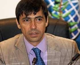 Un hombre vestido con un traje de color claro y corbata azul sentado en una mesa de conferencias con una expresión seria. Olive Oil Times