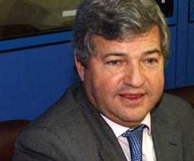 A man wearing a gray suit and blue tie seated at a table during a conference or meeting. - Olive Oil Times