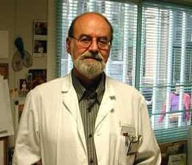 A man wearing a white lab coat stands in an office with a computer and various papers around. - Olive Oil Times