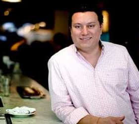 A man wearing a checkered shirt stands at a bar with a neutral expression. - Olive Oil Times