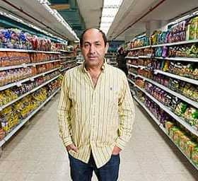 A man in a striped shirt standing in the aisle of a grocery store surrounded by various packaged products. - Olive Oil Times