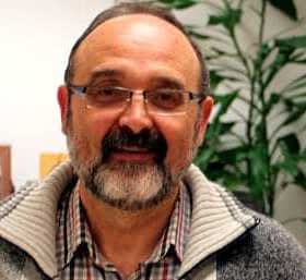 A man with glasses and a beard wearing a gray sweater, smiling in an indoor setting. - Olive Oil Times