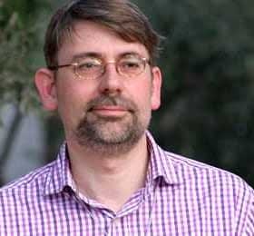 Man wearing a checked shirt with glasses, standing outdoors with a neutral expression. - Olive Oil Times