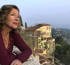 A woman with curly hair wearing a maroon top, looking out over a coastal landscape with buildings in the background. - Olive Oil Times