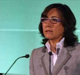 Woman with glasses speaking at a podium during a presentation with a green background. - Olive Oil Times