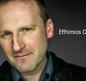 Close-up portrait of a man with short hair and a slight smile against a dark background. - Olive Oil Times