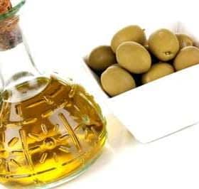 A glass bottle of olive oil with a cork stopper next to a bowl of green olives. - Olive Oil Times