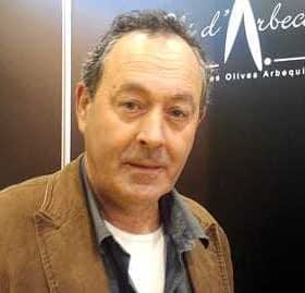 Homme aux cheveux foncés et à la chemise claire, portant une veste marron, debout devant un fond sombre. - Olive Oil Times