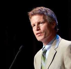 A man in a light suit and green tie speaking at a podium with a microphone. - Olive Oil Times