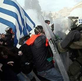 A protest scene in Greece featuring a person holding a Greek flag and police in riot gear. - Olive Oil Times
