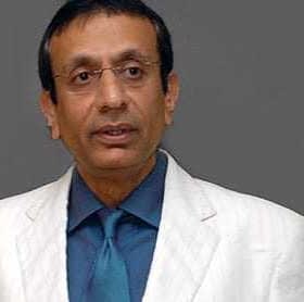 Man wearing a white suit with a blue shirt and tie, standing against a gray background. - Olive Oil Times