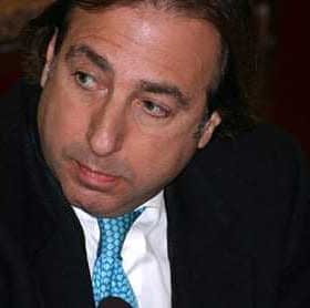 Man wearing a black suit and a blue tie, looking to the side during a discussion. - Olive Oil Times