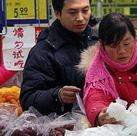 Deux individus sélectionnant des fruits sur un marché avec des étiquettes de prix visibles en arrière-plan. Olive Oil Times