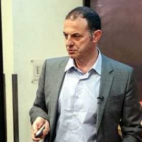 Un hombre vestido con un traje gris sostiene un puntero mientras realiza una presentación en un entorno interior. Olive Oil Times