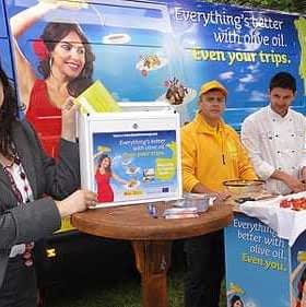 Woman holding a promotional box at an olive oil event with staff and a display table. - Olive Oil Times