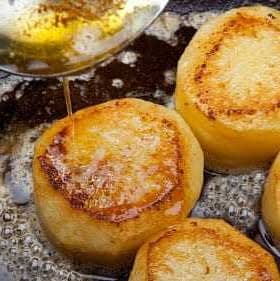 Golden-brown fried potato slices being drizzled with oil in a frying pan. - Olive Oil Times