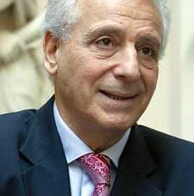 A man wearing a dark suit and a pink tie, smiling while seated indoors. - Olive Oil Times