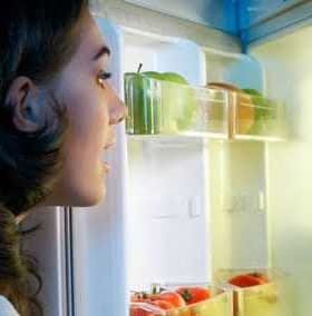 A person with dark hair looking into an open refrigerator filled with various food items and a bottle of oil. - Olive Oil Times