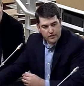 A man in a dark suit speaking during a meeting with two other men seated nearby. - Olive Oil Times