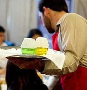 Waiter in a red apron holding a tray with two glasses of colored drinks at a dining event. - Olive Oil Times